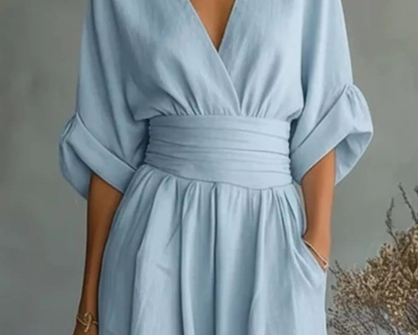 NEWYORKLISTED STYLE COLLECTION: A woman wearing a light blue midi dress with a wrap-style V-neckline, elbow-length puff sleeves, a matching fabric waist tie, and side pockets, photographed against a soft gray wall with dried floral decor.