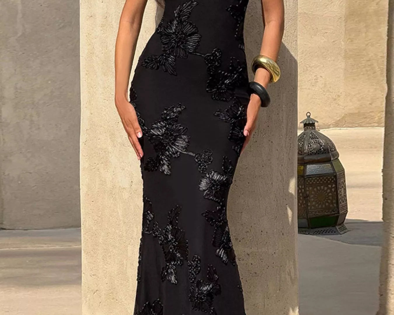 NEWYORKLISTED STYLE COLLECTION: Woman wearing a black evening gown with floral embroidery against a stone wall.