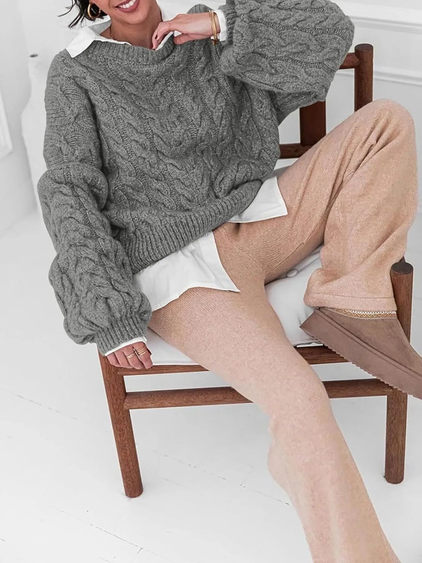 NEWYORKLISTED STYLE COLLECTION: Model wearing a gray cable knit sweater and beige pants sitting on a wooden chair.