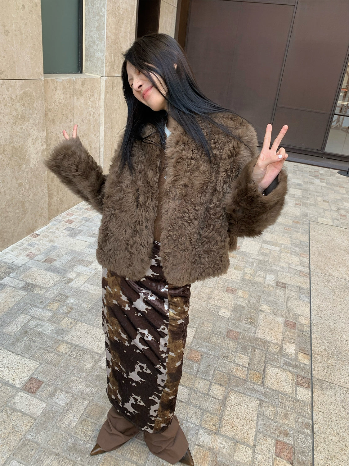 Person wearing a brown fur coat and patterned pants standing on a sidewalk.