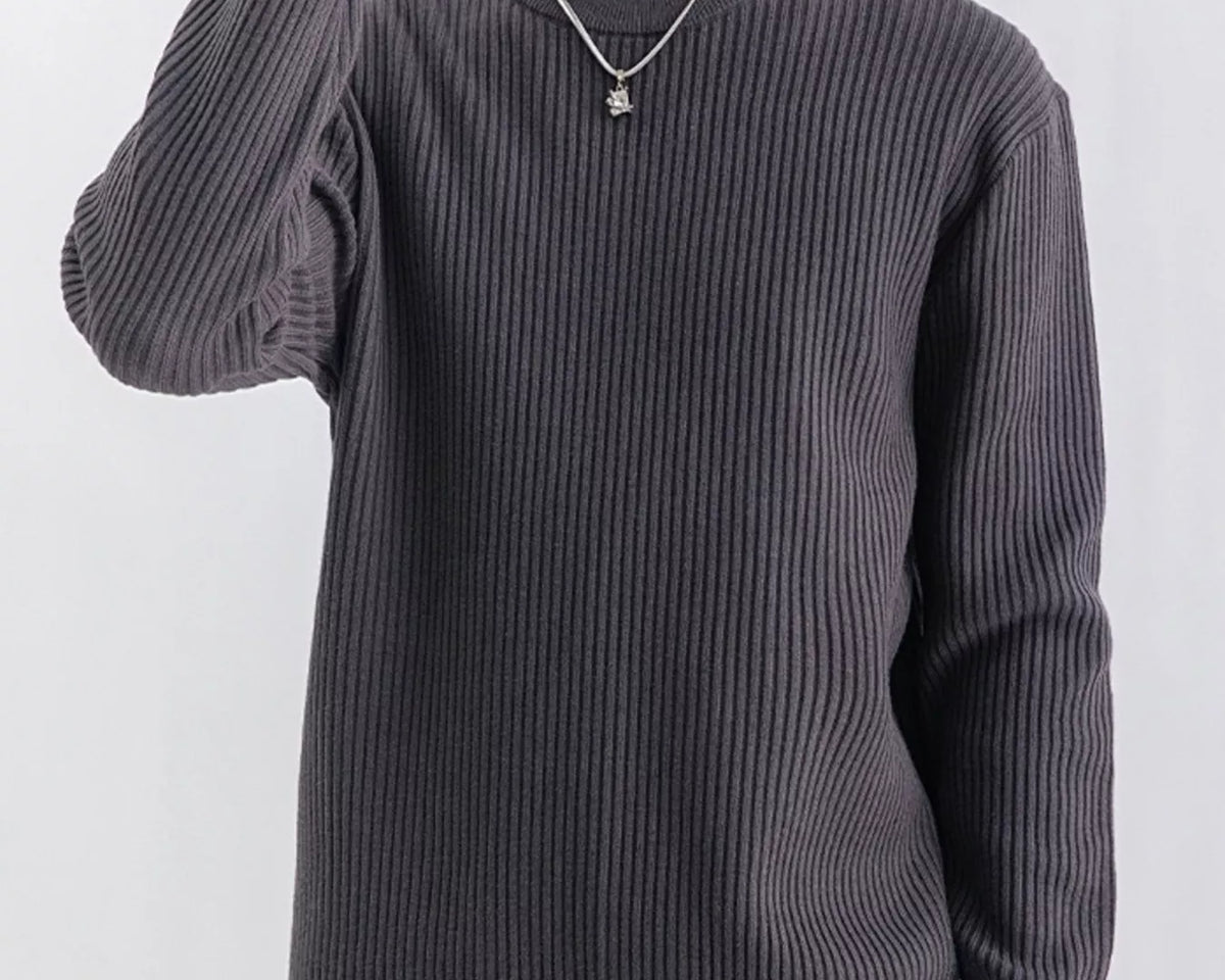 NEWYORKLISTED ATLAS MEN'S COLLECTION: A man wearing a dark gray ribbed knit crewneck sweater, paired with tan pants and a silver necklace, against a plain light gray background.