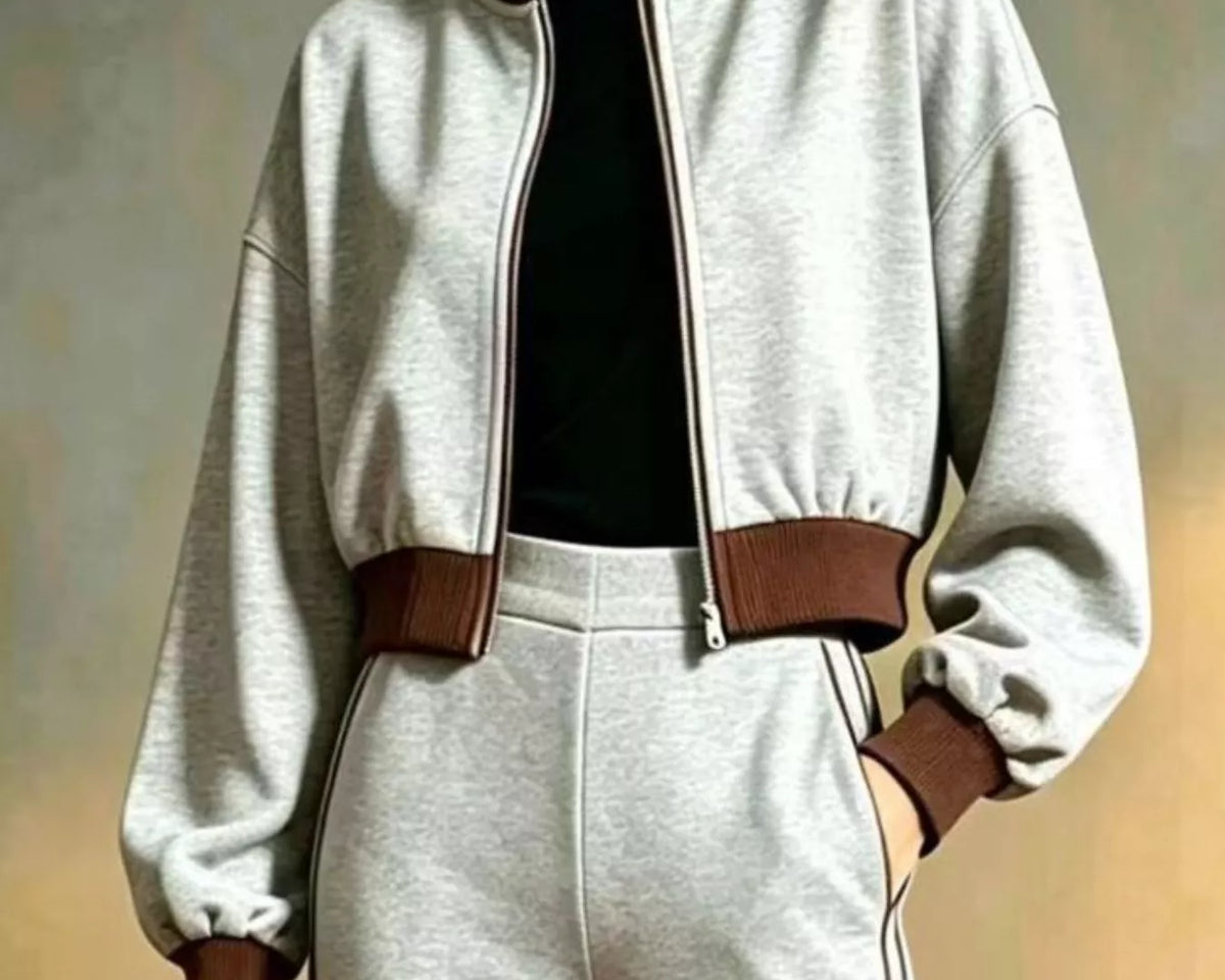 NEWYORKLISTED STYLE COLLECTION: A person wearing a light gray cropped zip-up jacket with brown ribbed collar, hem, and cuffs, paired with matching light gray sweatpants and a black top, against a soft neutral studio background.