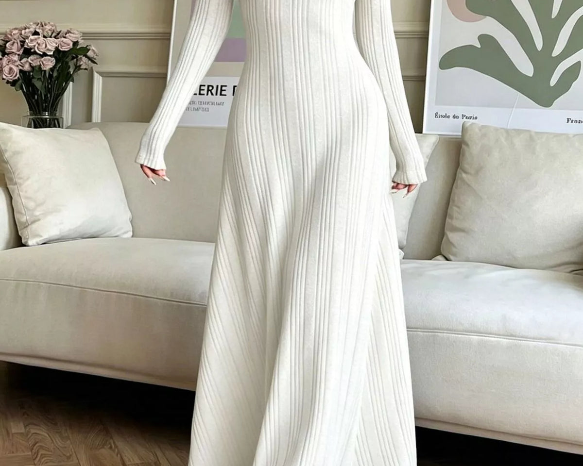 NEWYORKLISTED STYLE COLLECTION: A woman wearing a long-sleeve, floor-length white ribbed knit dress, standing in a living room setting with a beige sofa and framed wall art in the background.