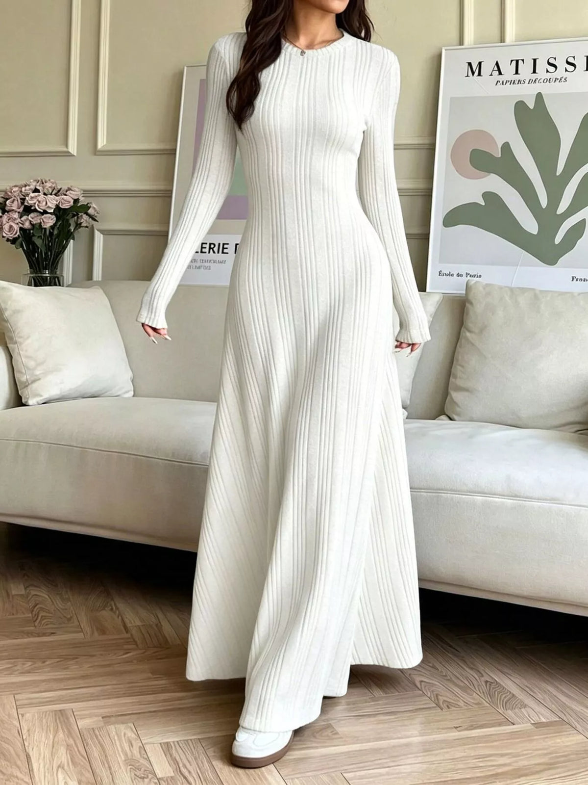 NEWYORKLISTED STYLE COLLECTION: A woman wearing a long-sleeve, floor-length white ribbed knit dress, standing in a living room setting with a beige sofa and framed wall art in the background.