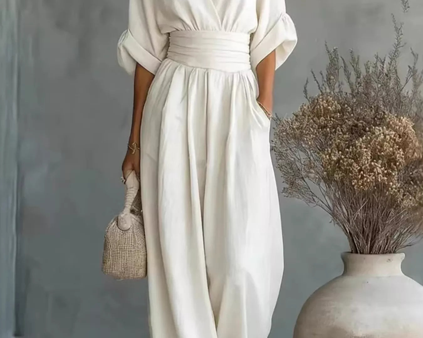 NEWYORKLISTED STYLE COLLECTION: Woman in a white dress standing next to a vase with dried flowers against a gray wall.
