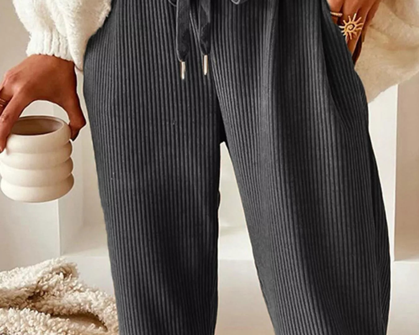 Person wearing dark ribbed pants with a white sweater and holding a ceramic cup.