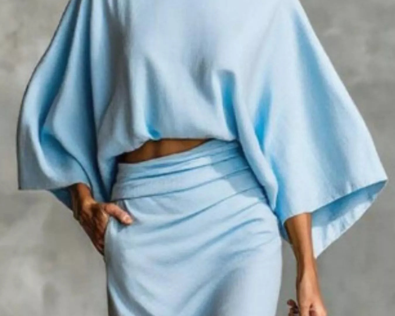 NEWYORKLISTED STYLE COLLECTION: Woman wearing a light blue outfit with a cape-like top against a gray background