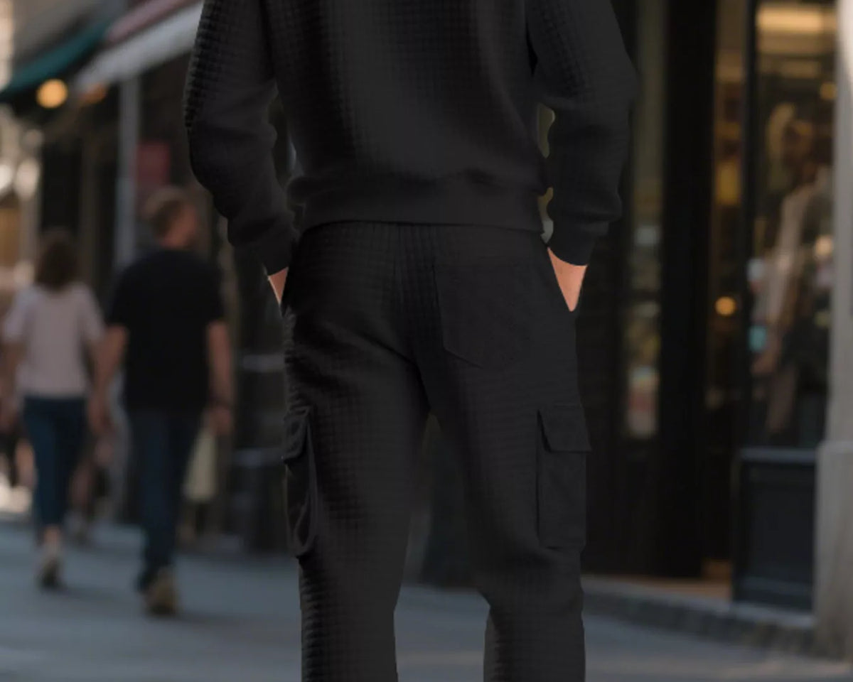 NEWYORKLISTED ATLAS MEN'S COLLECTION: A man viewed from behind wearing a matching black textured knit crewneck sweatshirt and cargo jogger pants on a city sidewalk.