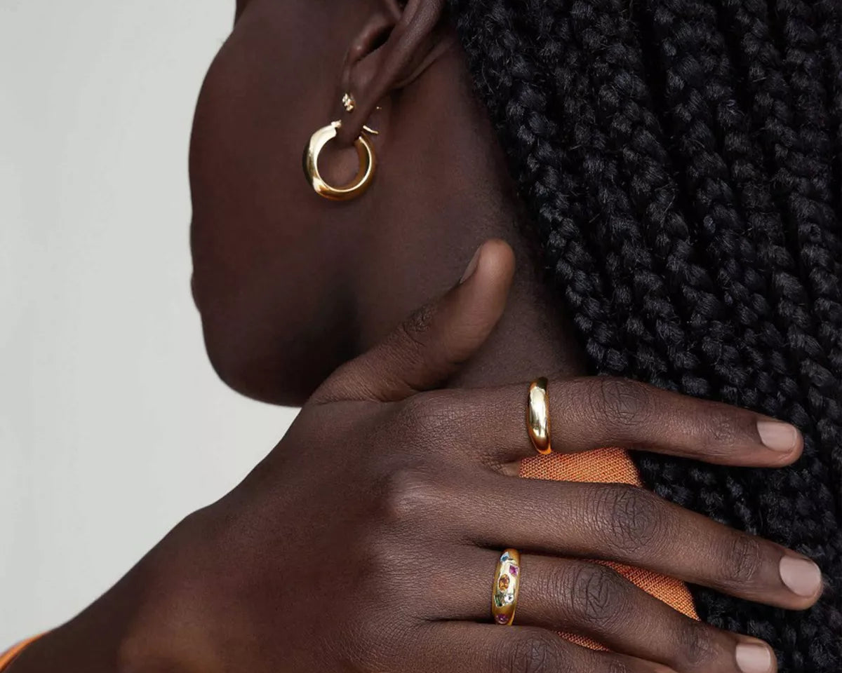 NYLD Glamour Collection by NEWYORKLISTED: Close-up of a person wearing gold hoop earrings and rings on a neutral background