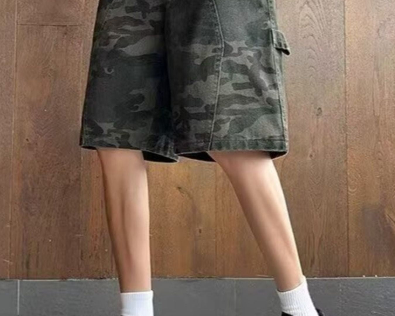 NEWYORKLISTED ATLAS MEN's COLLECTION: Person wearing camouflage shorts and black sneakers against a wooden wall.