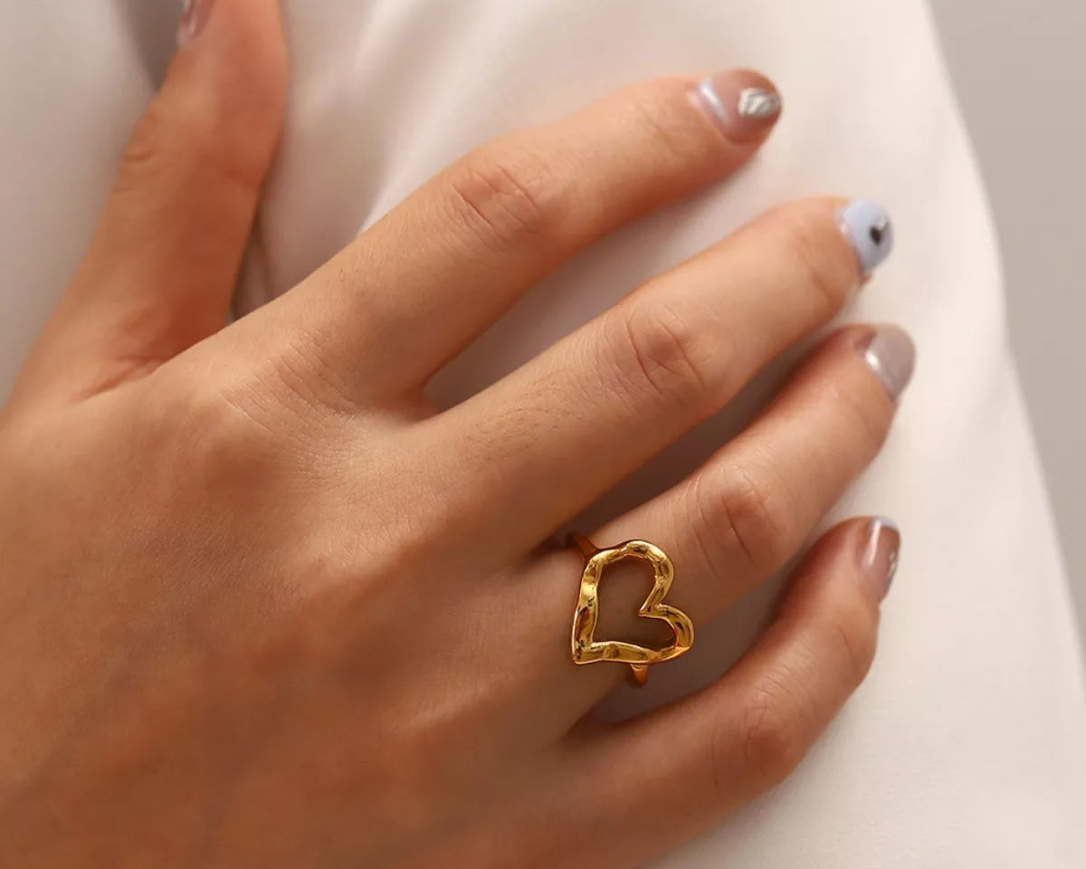 NYLD Glamour Collection by NEWYORKLISTED: Hand wearing a gold heart-shaped ring on a neutral background