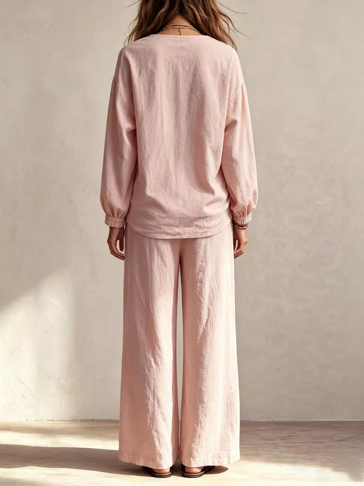 NEWYORKLISTED STYLE COLLECTION: A woman viewed from behind wearing a light pink linen two-piece set consisting of a long-sleeve top with gathered cuffs and matching wide-leg pants, standing against a textured beige wall.