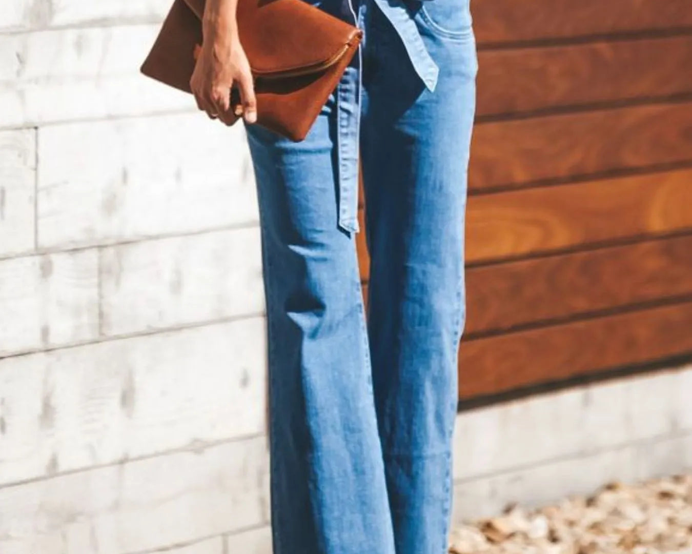NEWYORKLISTED STYLE COLLECTION: Person wearing blue jeans and a white top, holding a brown clutch against a wooden wall.
