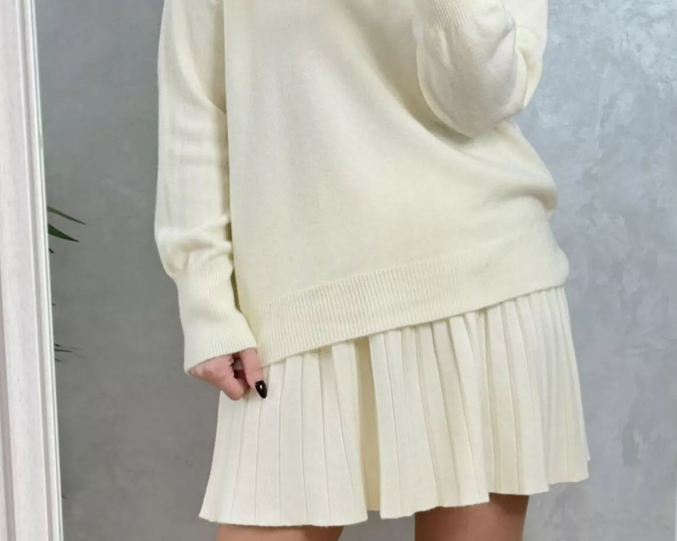 Person wearing a cream-colored sweater and pleated skirt with brown boots against a plain background on NEWYORKLISTED.COM