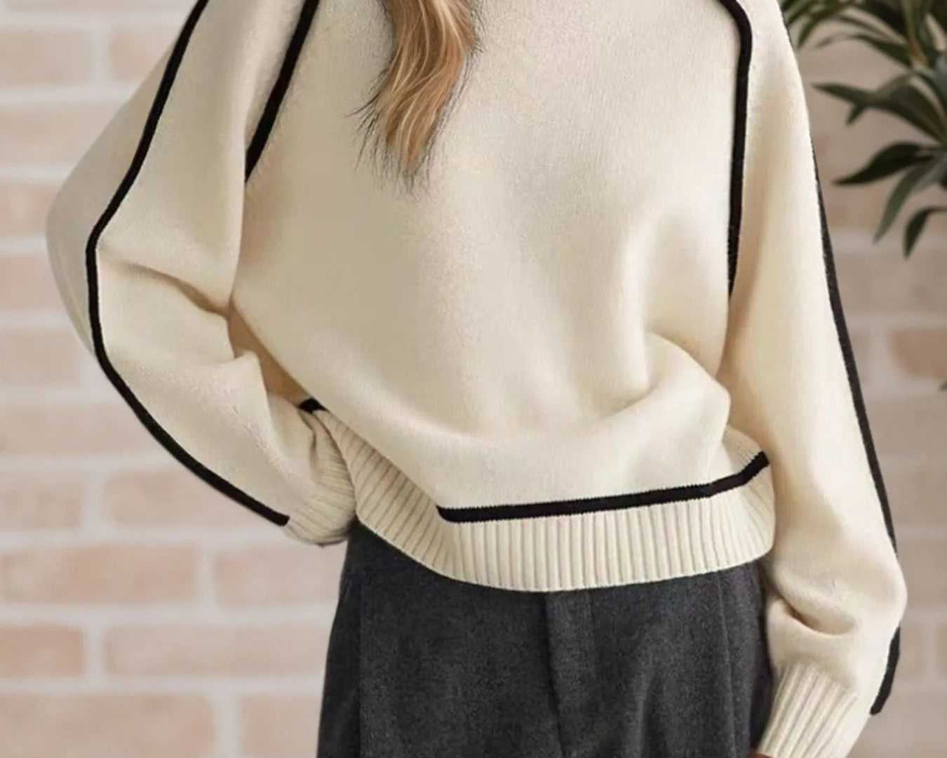 NEWYORKLISTE STYLE COLLECTION: Person wearing a cream sweater with black trim against a brick wall.