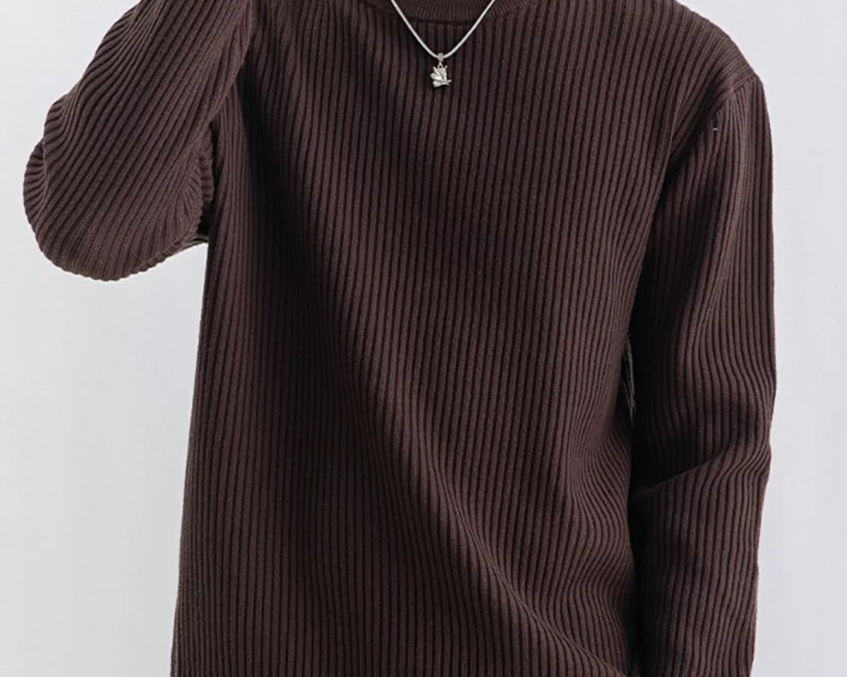 NEWYORKLISTED ATLAS MEN'S COLLECTION: A man wearing a dark brown ribbed knit crewneck sweater, paired with beige pants and a silver necklace, against a plain light gray background.