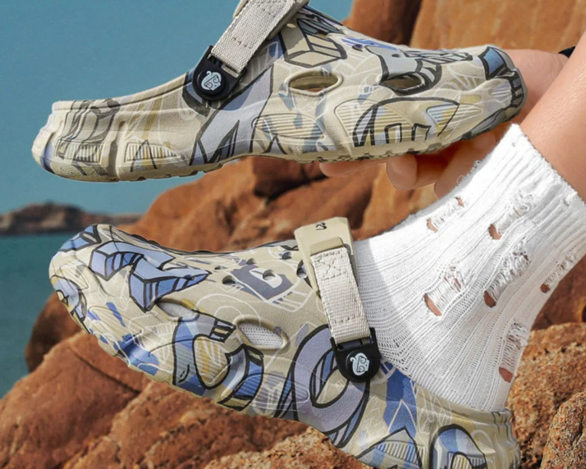 NEWYORKLISTED ATLAS MEN'S COLLECTION: Pair of patterned sandals held by a person on rocky terrain with water in the background