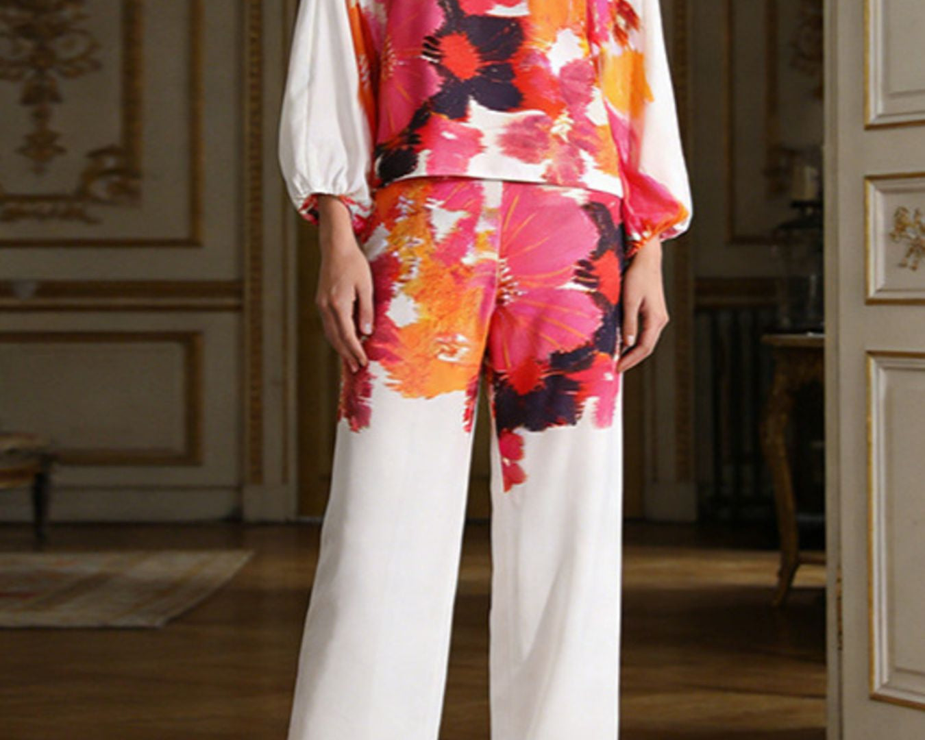 NEWYORKLSITED STYLE COLLECTION: Woman wearing a colorful floral off-shoulder top and white pants in an elegant room.