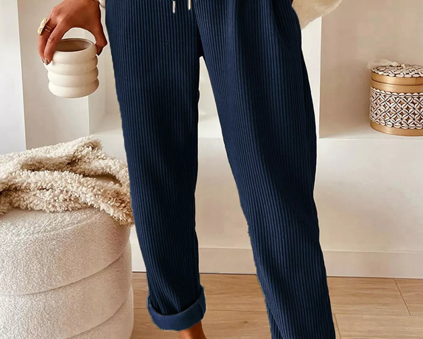 NEWYORKLISTED STYLE COLLECTION: Person wearing navy ribbed pants with a white sweater and brown shoes, holding a small white container.
