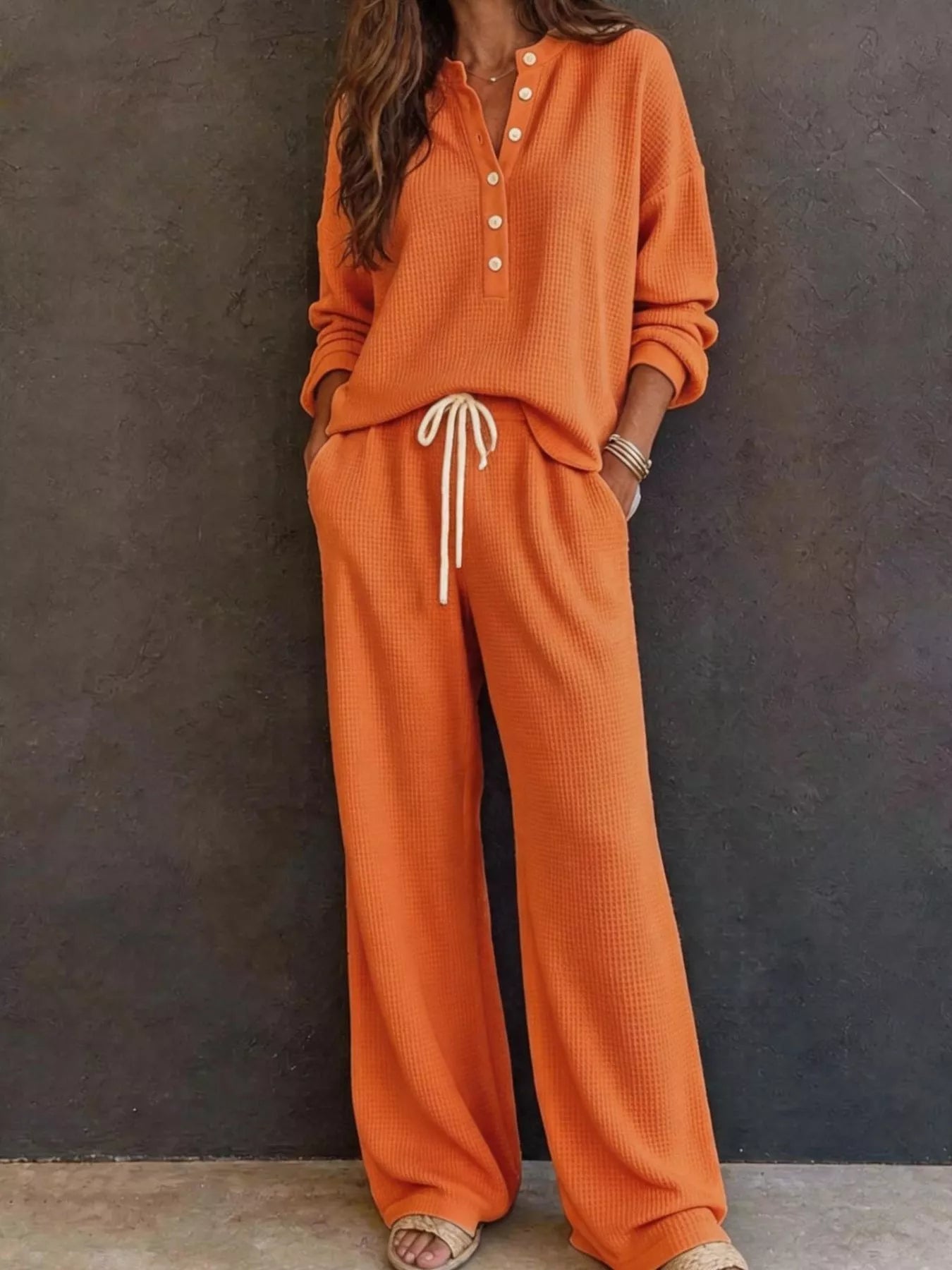 NEWYORKLISTED STYLE & COMFORT COLLECTION: A woman wearing a matching orange waffle-knit lounge set with a long-sleeve henley top and wide-leg pants, featuring a white drawstring, standing against a dark textured wall.
