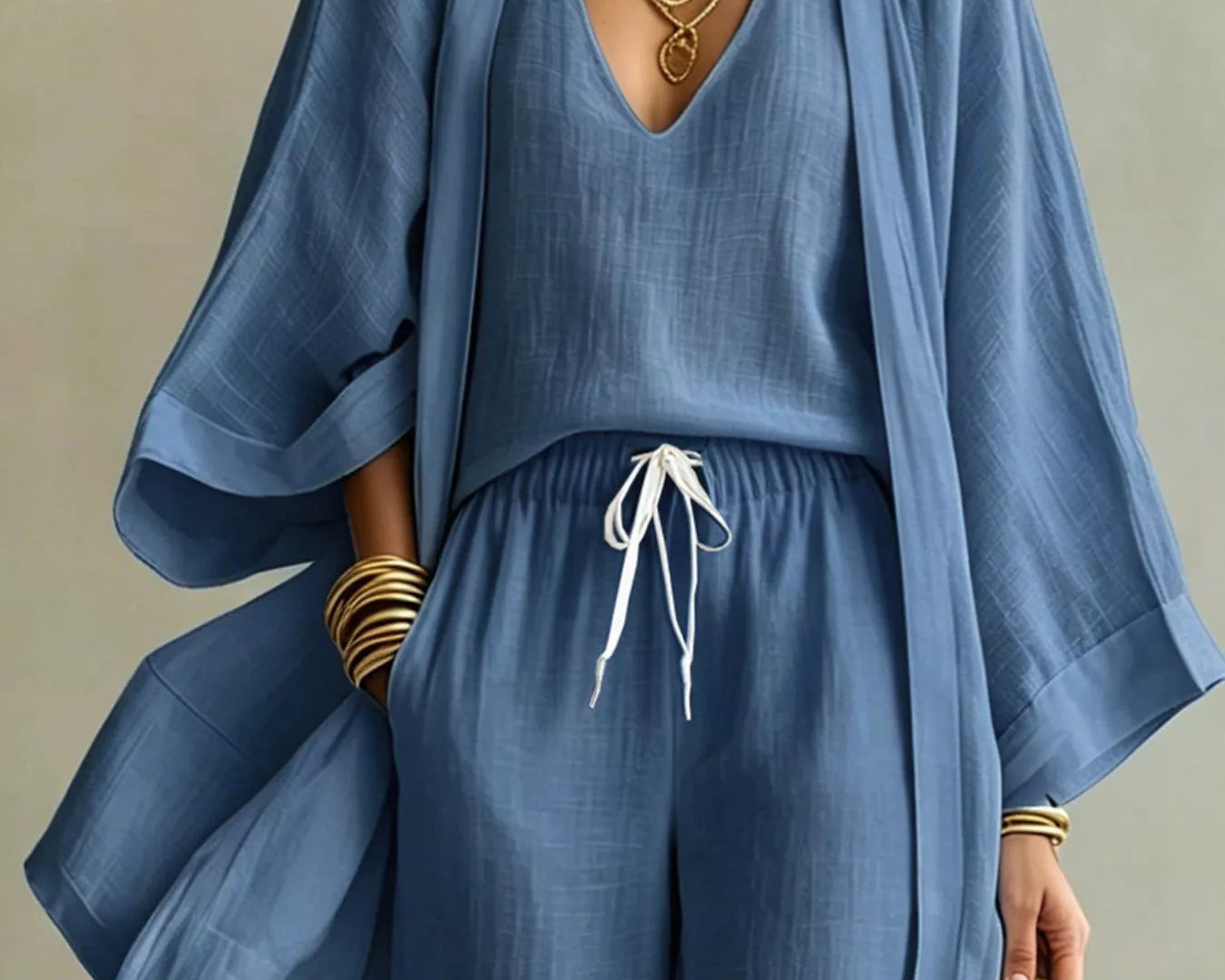A woman wearing a matching blue NEWYORKLISTED STYLE COLLECTION: two-piece outfit with a loose long-sleeve top and wide-leg pants, styled with layered gold necklaces, bracelets, and a woven handbag against a neutral background.