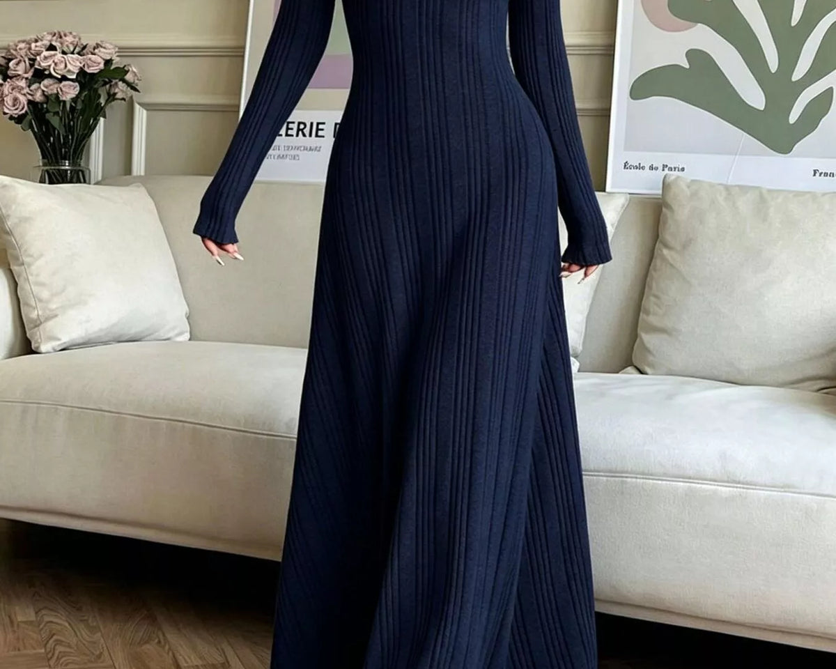 NEWYORKLISTED STYLE COLLECTION: A woman wearing a navy blue long-sleeve maxi dress with a vertical pleated texture, standing in a living room setting with a beige sofa and wooden floor.