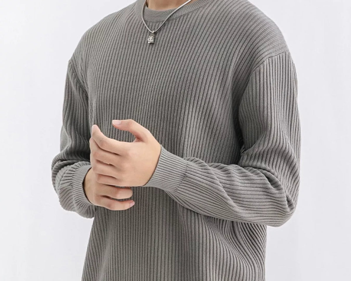 NEWYORKLISTED ATLAS MEN'S COLLECTION: A man wearing a ribbed gray long-sleeve crewneck sweater, paired with tan pants and a silver necklace, photographed against a clean white studio background.
