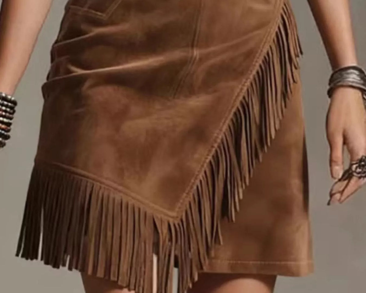 NEWYORKLISTED STYLE COLLECTION: A brown suede mini skirt with asymmetrical fringe detailing, shown on a model from the waist down against a neutral gray background.