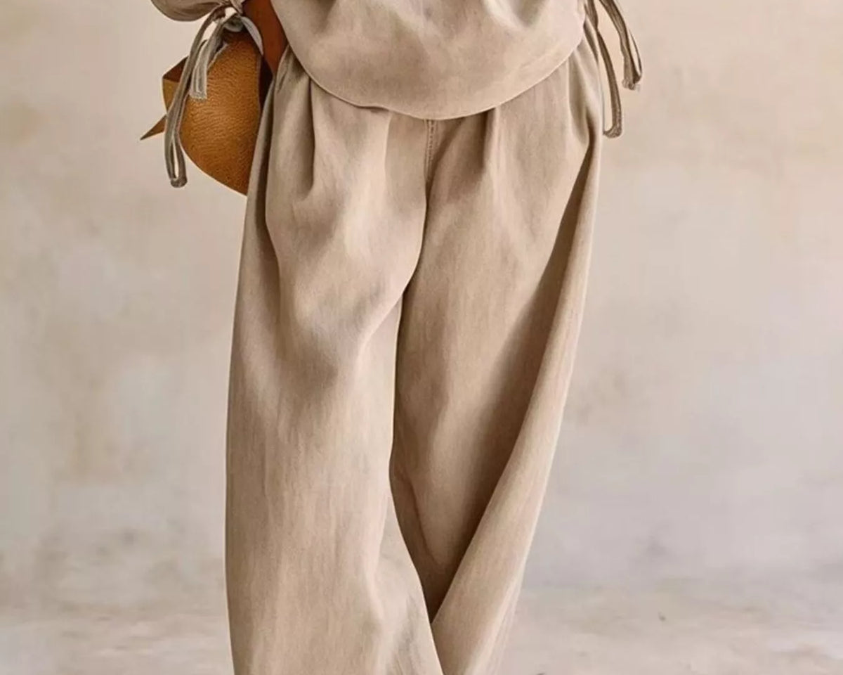 NEWYORKLISTED STYLE COLLECTION: A person wearing a matching beige linen-like two-piece outfit with a loose long-sleeve top and wide-leg pants, styled with a small round tan bag and white sneakers against a soft neutral wall.