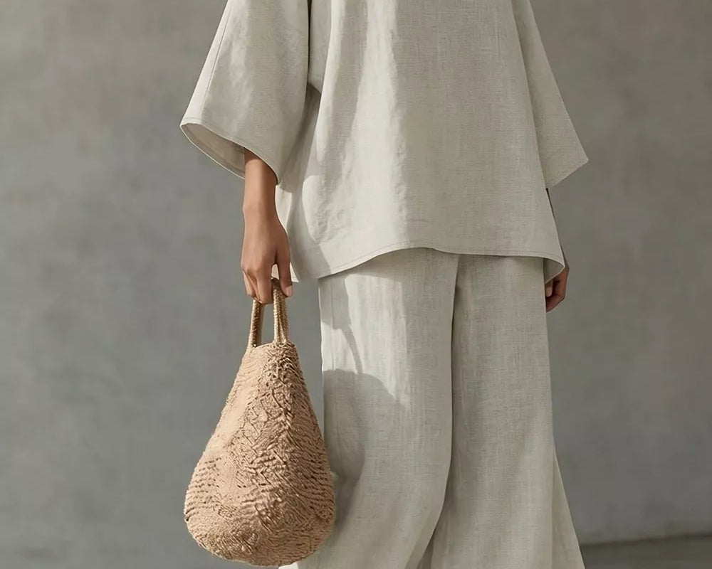 NEWYORKLISTED STYLE COLLECTION: A model wearing a light beige linen two-piece set consisting of a loose-fitting short-sleeve top and matching wide-leg pants, holding a woven straw handbag against a textured gray wall.