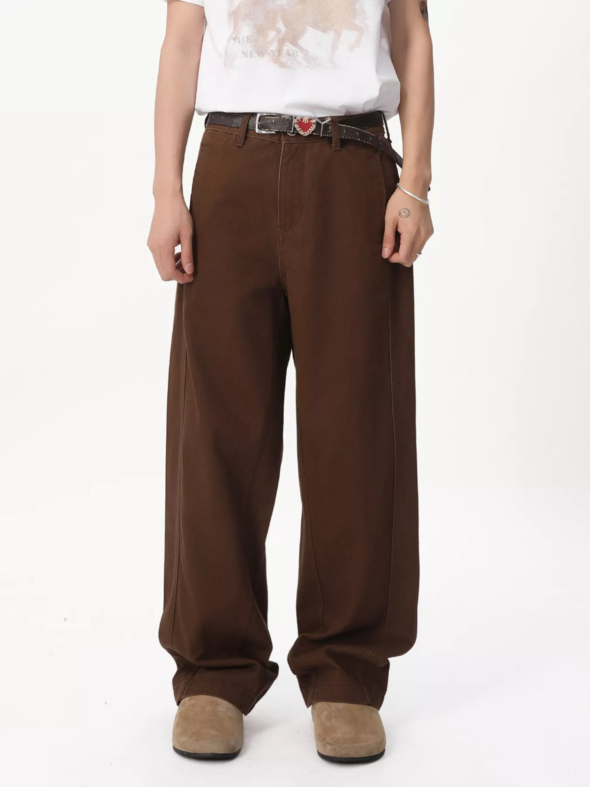 NEWYORKLISTED ATLAS MEN'S COLLECTION: A person wearing loose brown wide-leg pants with a dark belt and a small red heart-shaped buckle detail, paired with a white graphic T-shirt and tan shoes, photographed against a plain white studio background.