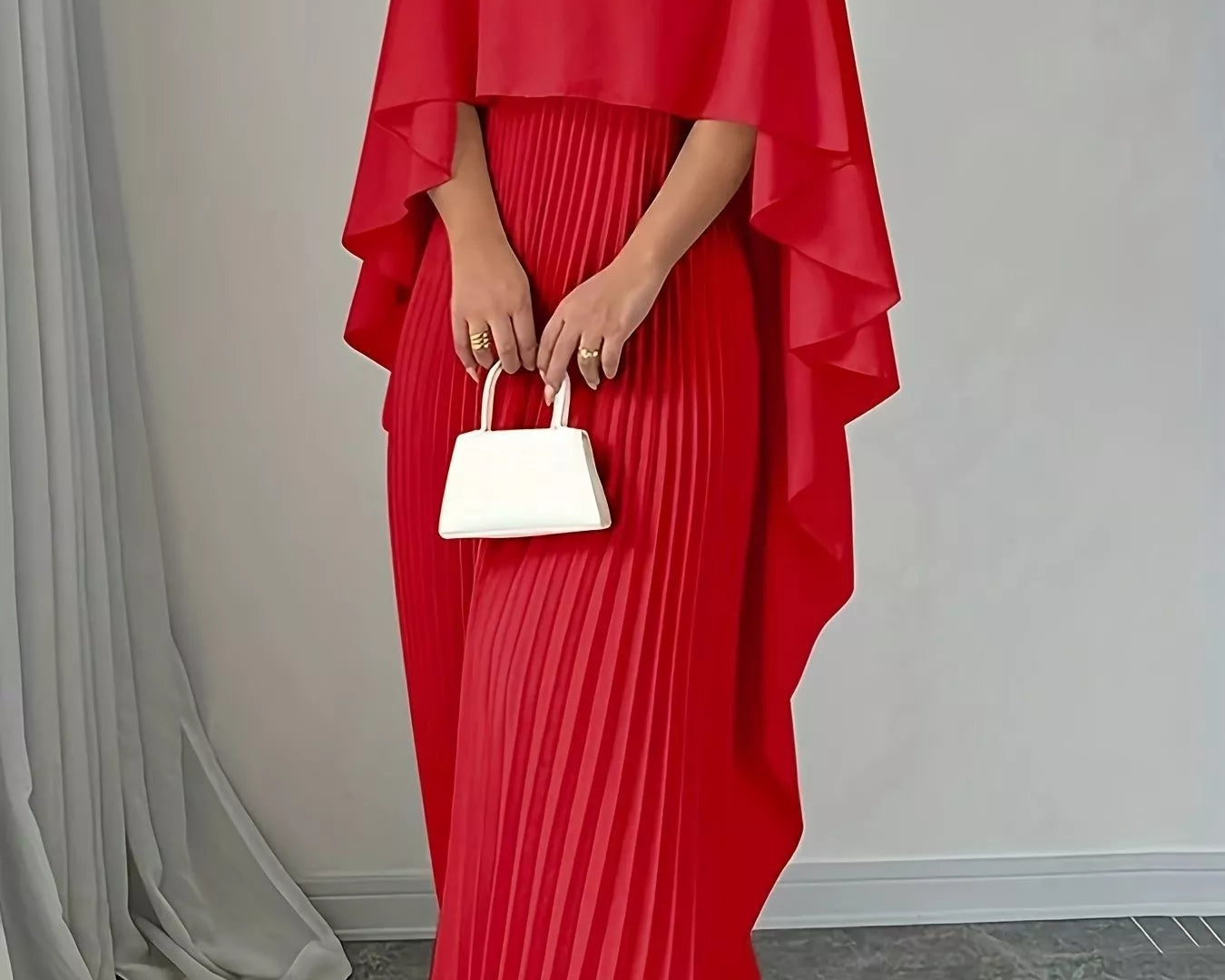 NEWYORKLISTED STYLE COLLECTION: Red pleated dress with cape held by a person against a neutral background