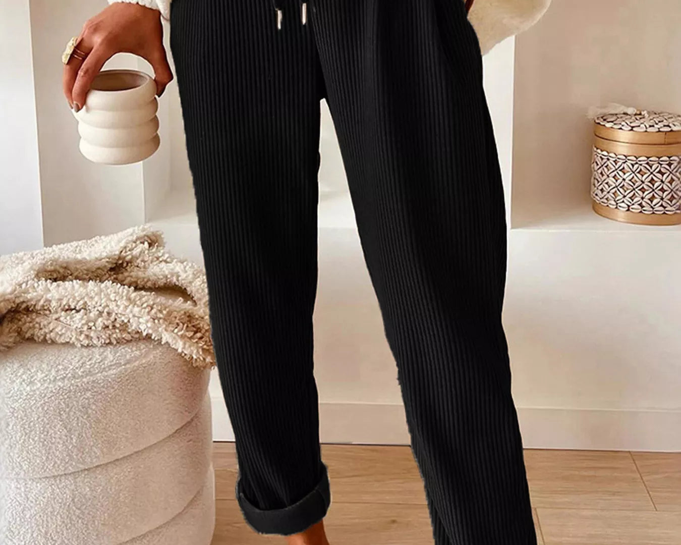 NEWYORKLISTED STYLE COLLECTION: Person wearing black ribbed pants and a white sweater, holding a ceramic cup indoors.
