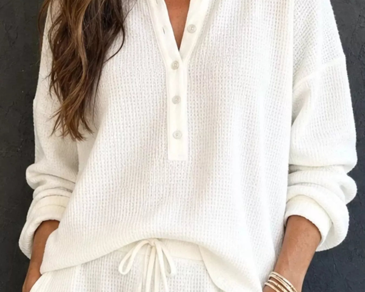 NEWYORKLISTED STYLE & COMFORT COLLECTION: A woman wearing a cream-colored waffle-knit long-sleeve henley top and matching drawstring pants, standing against a dark textured wall.