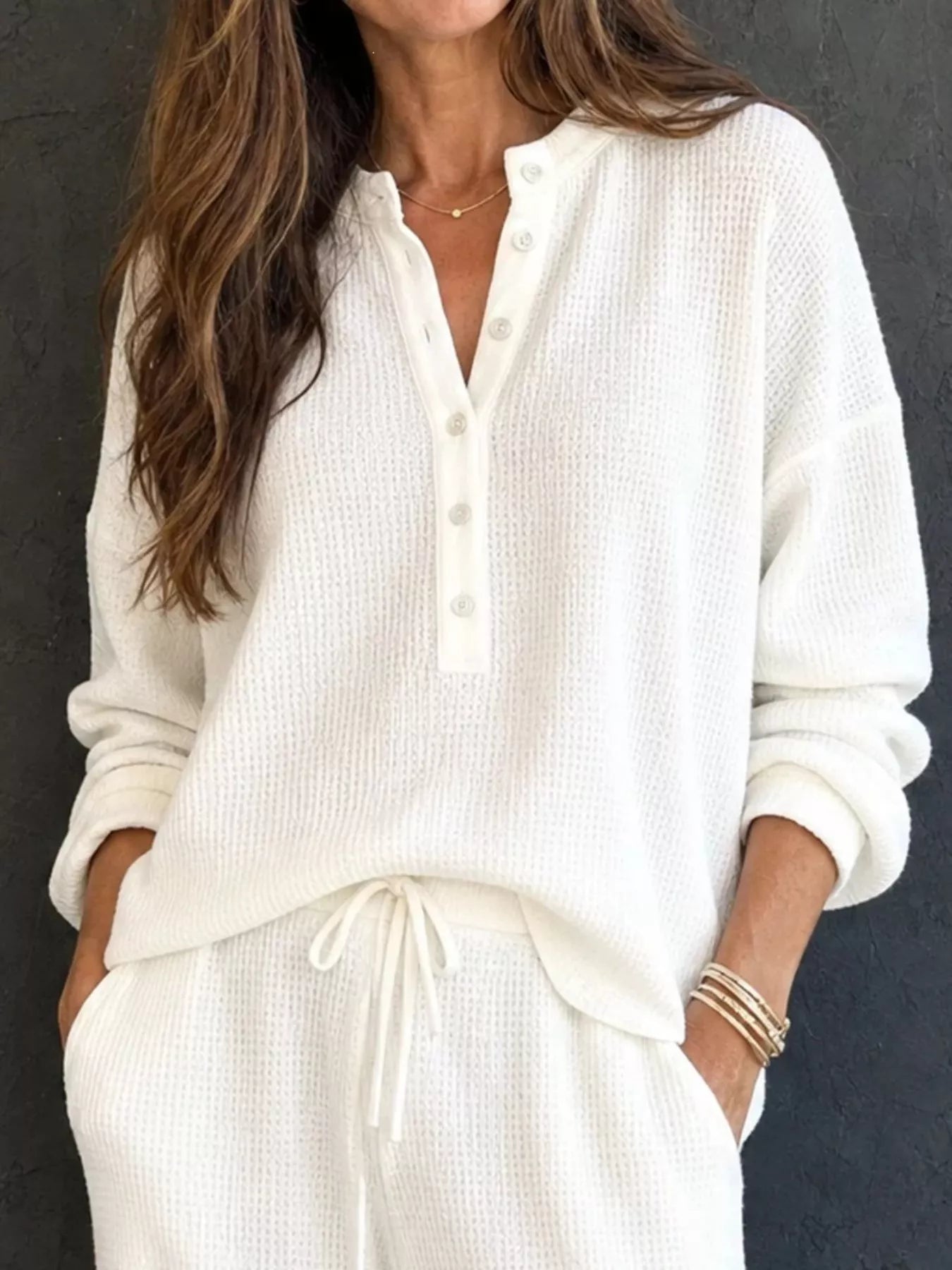 NEWYORKLISTED STYLE & COMFORT COLLECTION: A woman wearing a cream-colored waffle-knit long-sleeve henley top and matching drawstring pants, standing against a dark textured wall.