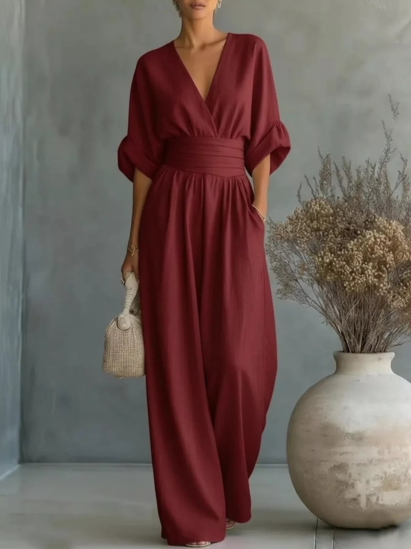 NEWYORKLISTED STYLE COLLECTION: Woman wearing a burgundy jumpsuit holding a woven bag against a gray wall with a vase of dried flowers.