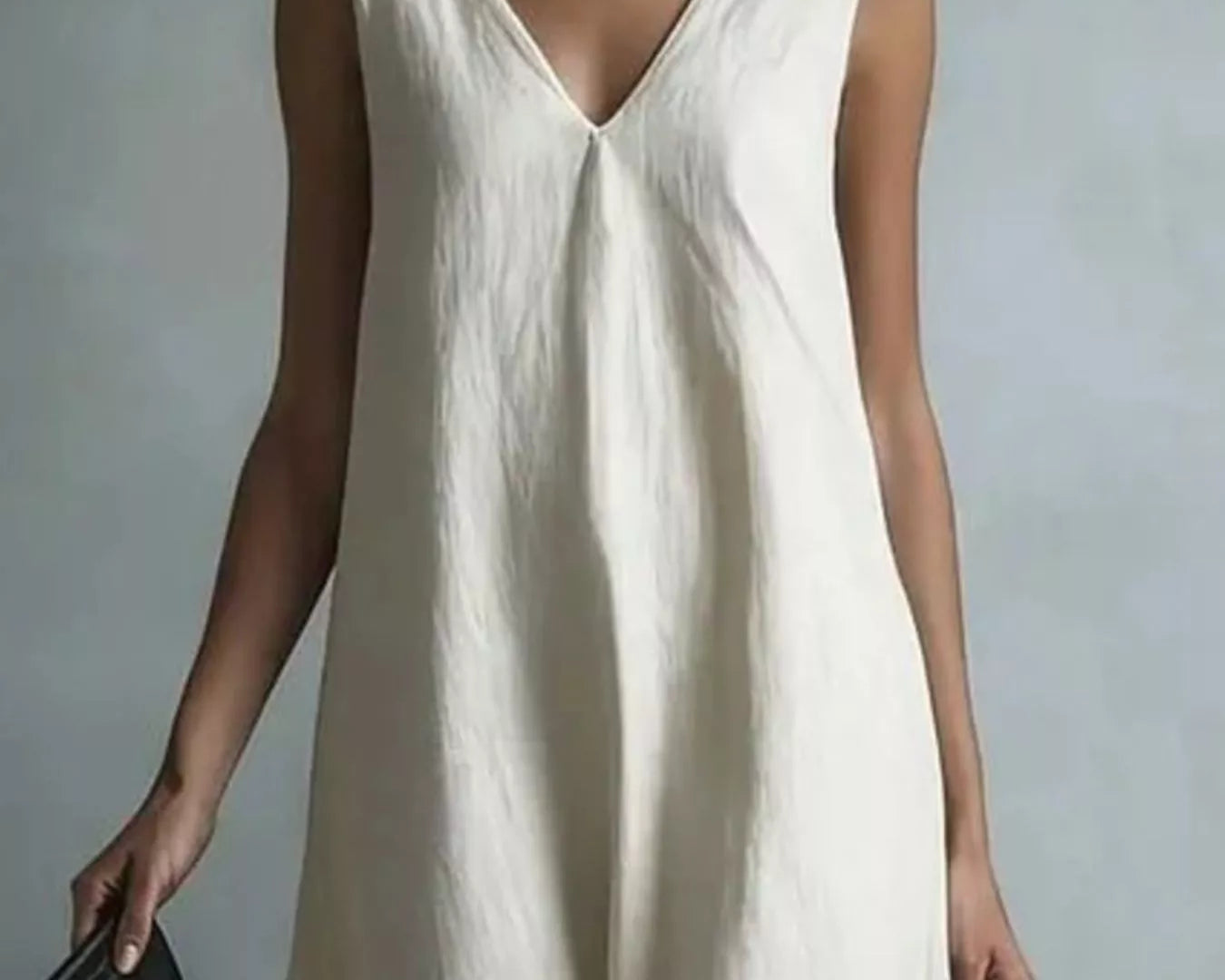 NEWYORKLISTED STYLE COLLECTION: A woman wearing a sleeveless, V-neck cream linen jumpsuit, holding a black leather clutch against a neutral gray background.