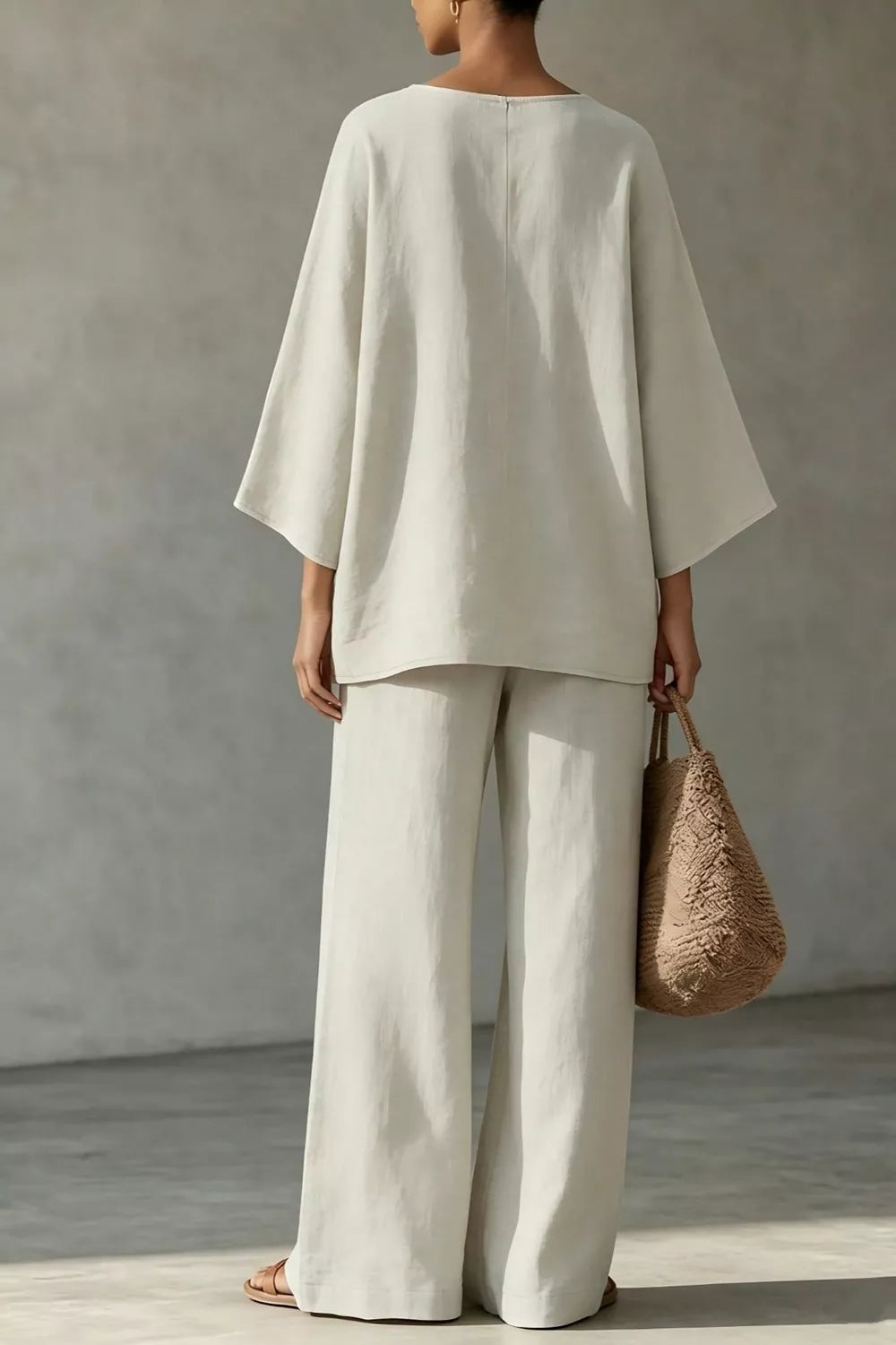NEWYORKLISTED STYLE COLLECTION: A woman shown from the back wearing a loose off-white linen two-piece set with a wide-sleeve top and wide-leg trousers, holding a woven straw bag against a textured gray wall.