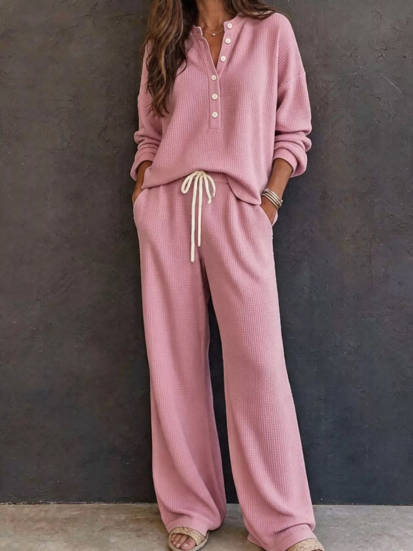 NEWYORKLISTED STYLE & COMFORT COLLECTION: A woman wearing a matching dusty pink lounge set consisting of a long-sleeve button-front top and wide-leg pants with a cream drawstring, standing against a dark textured wall.
