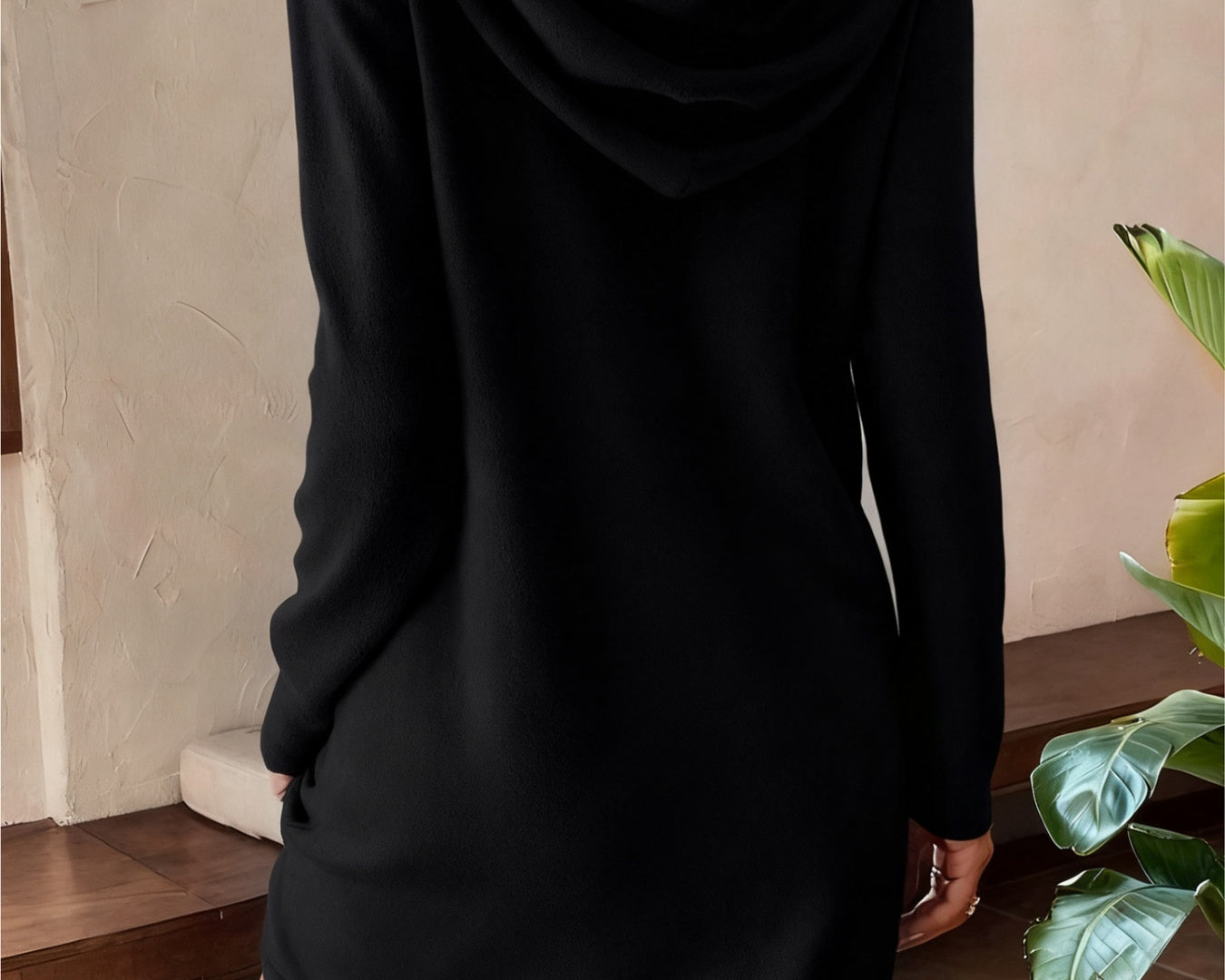 NEWYORKLISTED Comfort Collection: Person wearing a black hooded dress in a room with a plant.