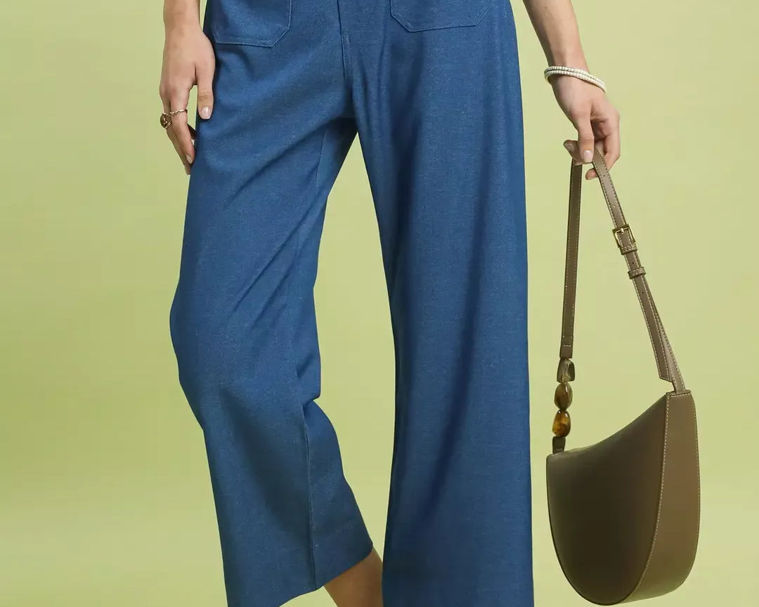 NEWYORKLISTED Style Collection: Person wearing blue pants and a light blue shirt, holding a brown handbag on a green background.