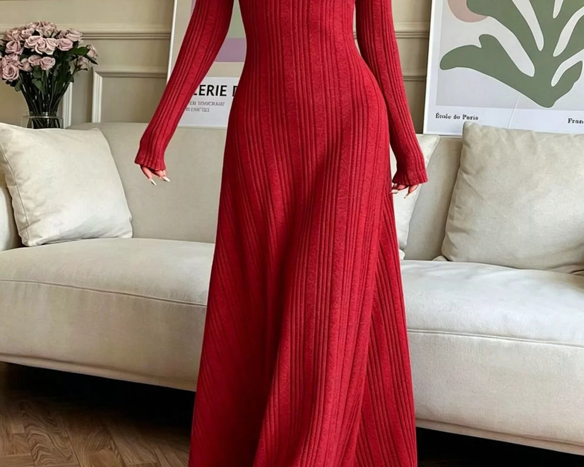 NEWYORKLISTED STYLE COLLECTION: A woman wearing a long-sleeved, floor-length red ribbed knit maxi dress, standing in a living room setting with a beige sofa and framed art in the background.