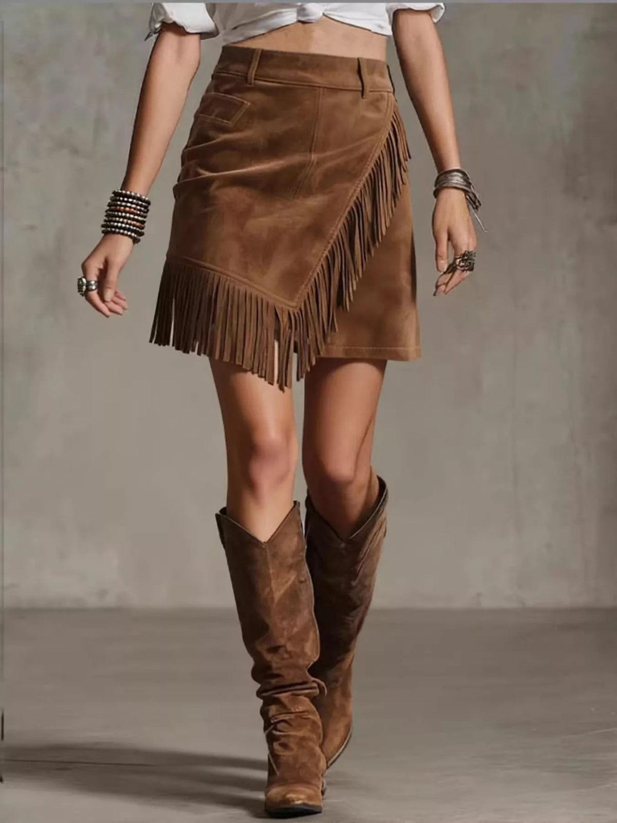 NEWYORKLISTED STYLE COLLECTION: A woman wearing a brown suede fringe mini skirt with an asymmetrical wrap design, paired with matching brown suede knee-high boots and silver bracelets, set against a textured gray studio background.