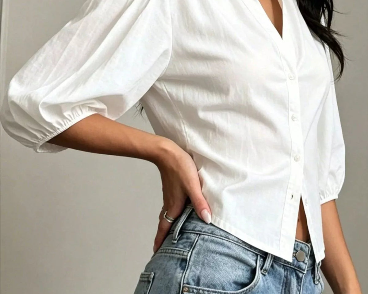 NEWYORKLISTED STYLE COLLECTION: A woman wearing a white short puff-sleeve button-up shirt tucked into light-wash blue jeans, shown from the neck down against a neutral indoor wall.