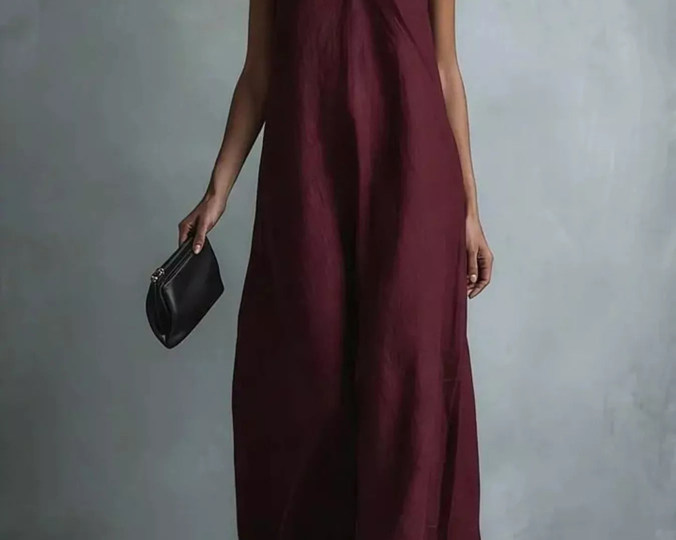 NEWYORKLISTED STYLE COLLECTION: A woman models a sleeveless, deep burgundy maxi dress with a V-neckline, holding a small black clutch against a textured gray studio backdrop.
