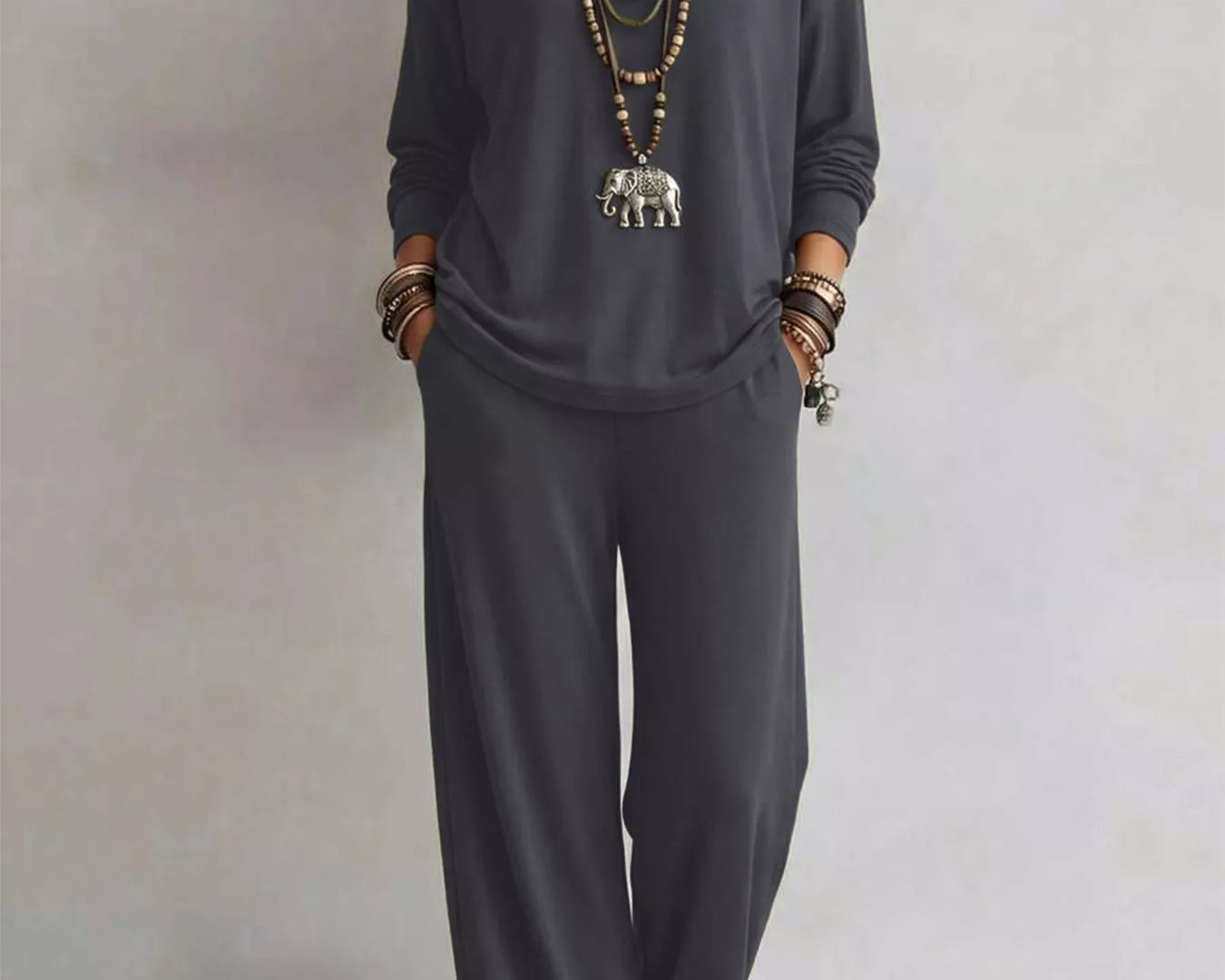 NEWYORKLISTED STYLE COLLECTION: A woman modeling a dark gray two-piece lounge set consisting of a long-sleeve top and matching wide-leg pants, styled with layered gold necklaces and bracelets against a neutral studio background.