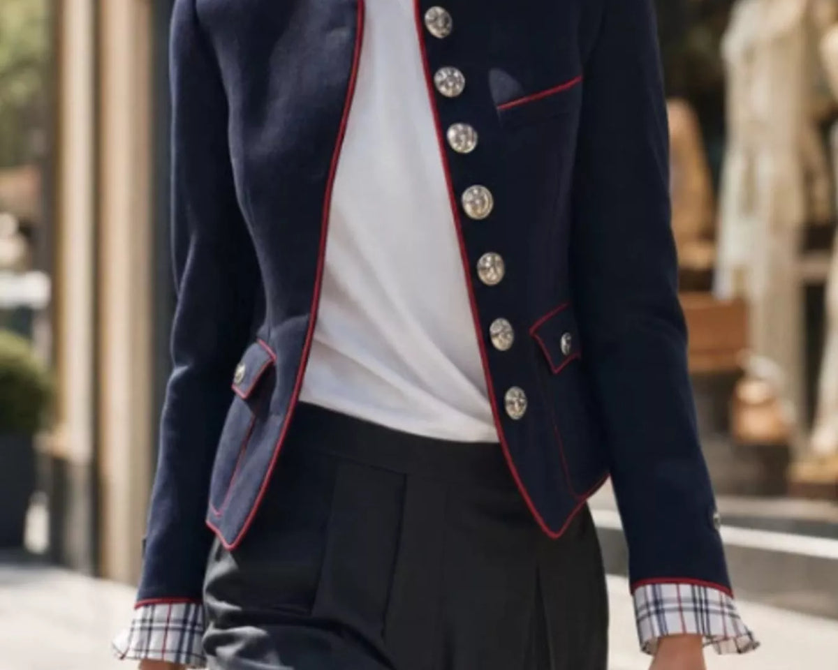 NEWYORKLISTED STYLE COLLECTION: A woman walking outdoors wearing a navy blazer with red piping and silver buttons, a white top, dark trousers, and sunglasses, holding a shopping bag.