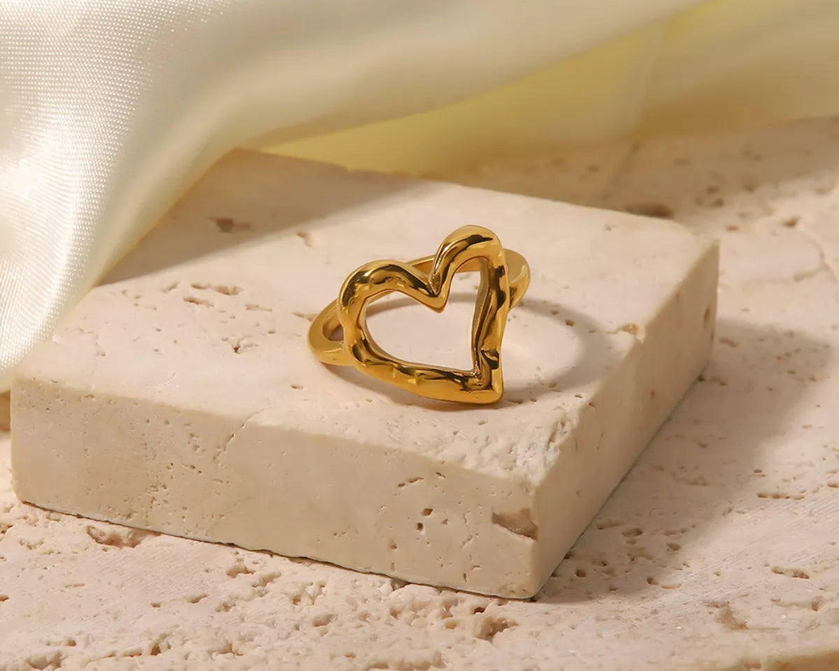 NYLD Glamour Collection by NEWYORKLISTED: Gold heart-shaped ring on a beige stone surface with a soft fabric background