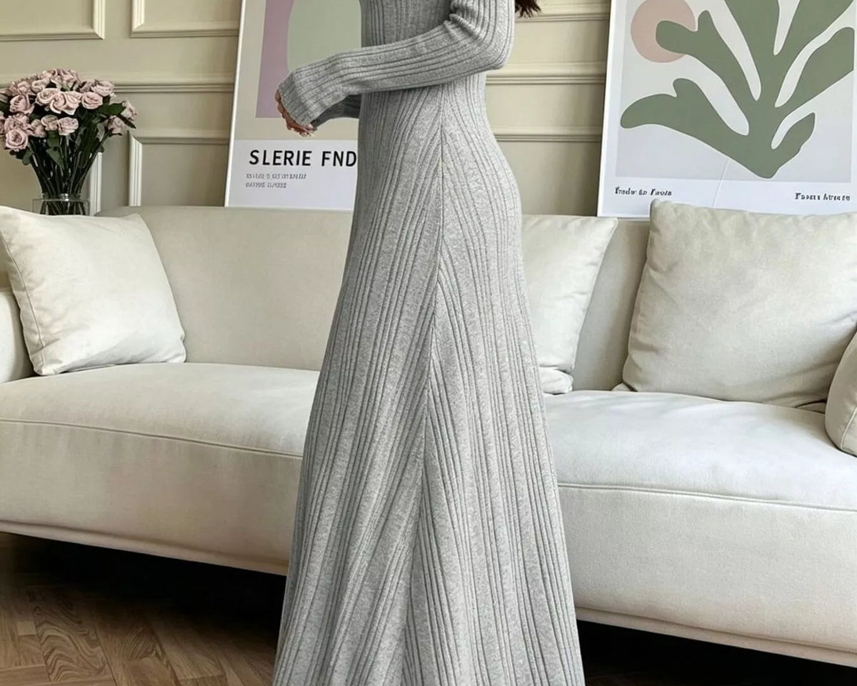 NEWYORKLISTED STYLE COLLECTION: A woman models a light gray ribbed knit maxi dress with long sleeves and a V-neckline in a bright, modern living room setting.
