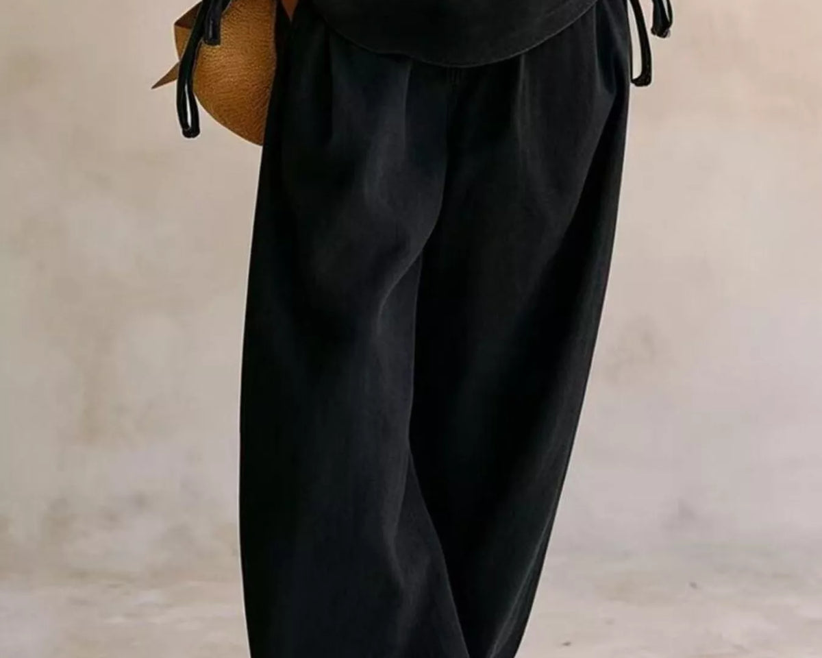 NEWYORKLISTED STYLE COLLECTION: A person wearing loose black drawstring pants and a matching black top, holding a small tan woven bag, photographed against a soft beige studio backdrop.